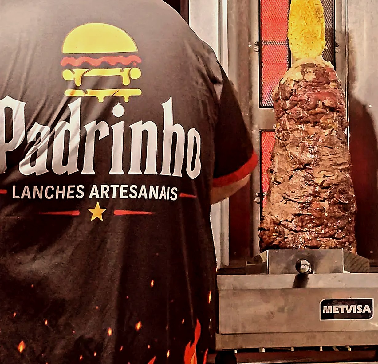 Atendente do Padrinho, de costas, ao lado do espeto giratório com a carne assando